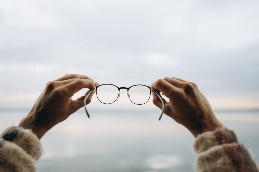 two hands hold a pair of glasses in front of an out of focus water scene in muted tones