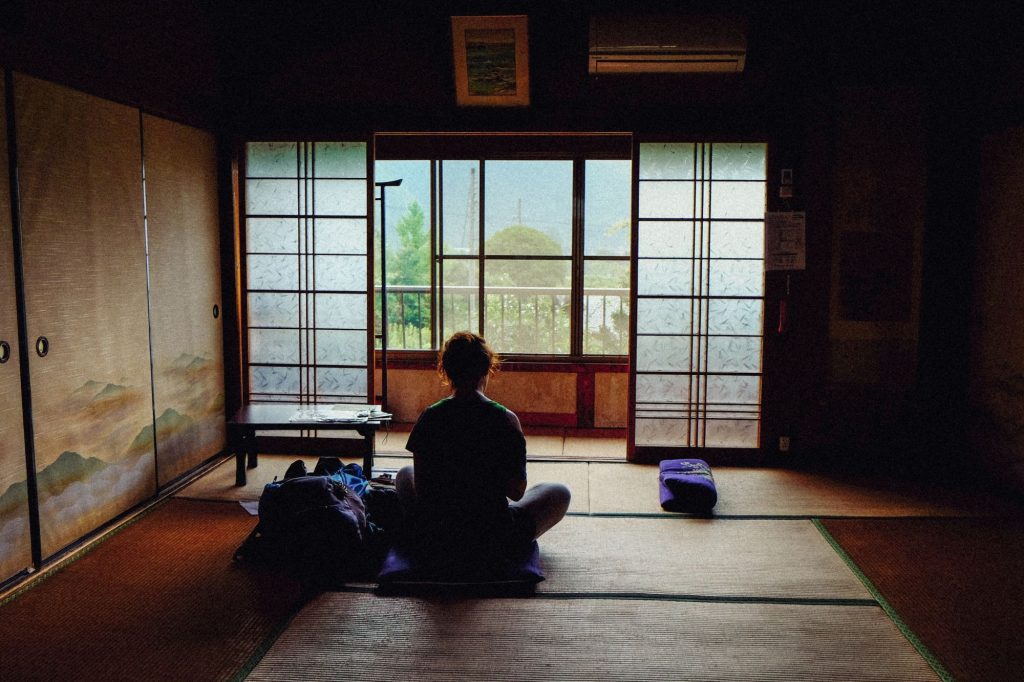 Someone is sitting on the floor in shadows, possibly meditating, in simple room which appears japanese