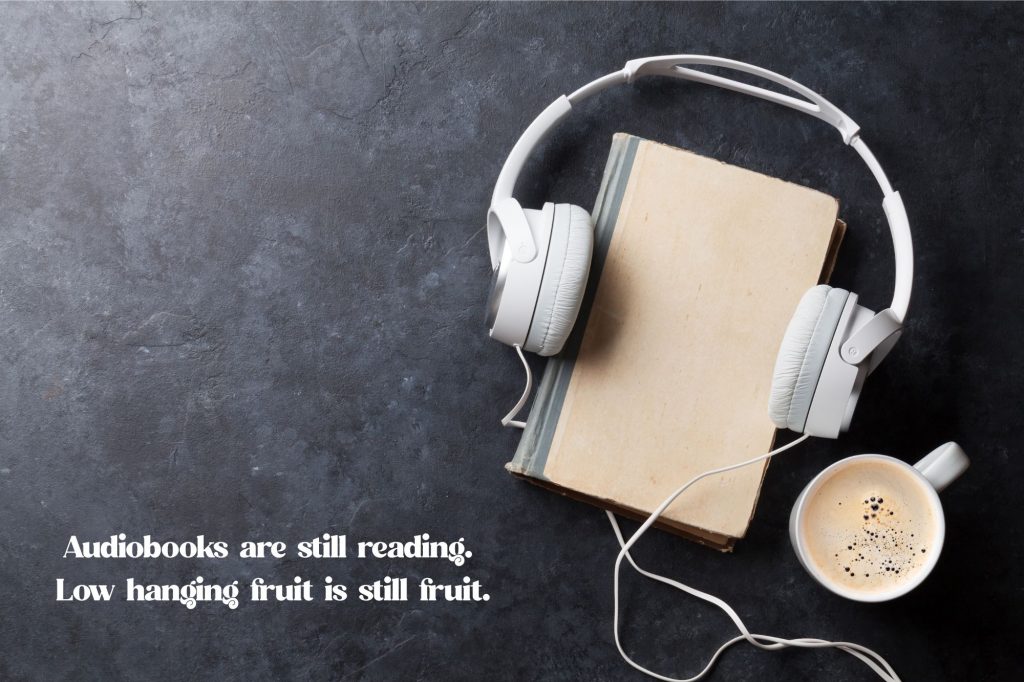 a book is on a slate background with headphones on either said and a cup of coffee next to it with a text overlay that reads "Audiobooks are still reading. Low hanging fruit is still fruit."
