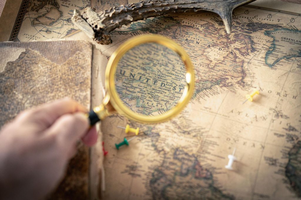 Out of focus hand holds a magnifying glass over a map with an antler on it, and thumbtacks strewn about. The area in the magnifying glass is clear 