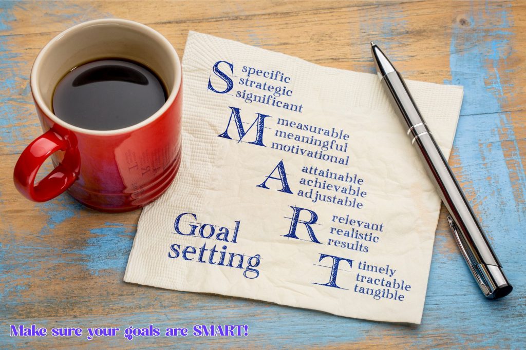 A coffee cup sits on a napkin with a pen on top. The napkin has the word "SMART" written on it as well as the phrase "goal setting," and the SMART is an acronym. The S shows "Specific, strategic, significant" the M shows "measurable, meaningful, motivational" and the A shows "Attainable, achievable, adjustable" and the R shows "relevant, realistic, results" and the T has "timely, tractable, tangible." Then on the bottom there's a text overlay that reads "make sure your goals are SMART!" 