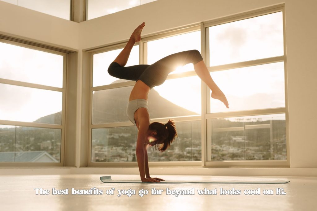 a woman does a handstand with legs spread artistically in front of a huge window and sunshine with text overlay that reads "the best benefits of yoga go far beyond what looks cool on IG." 