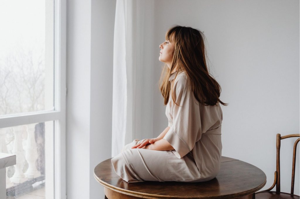 A woman sits cross-legged on. a table looking out a window