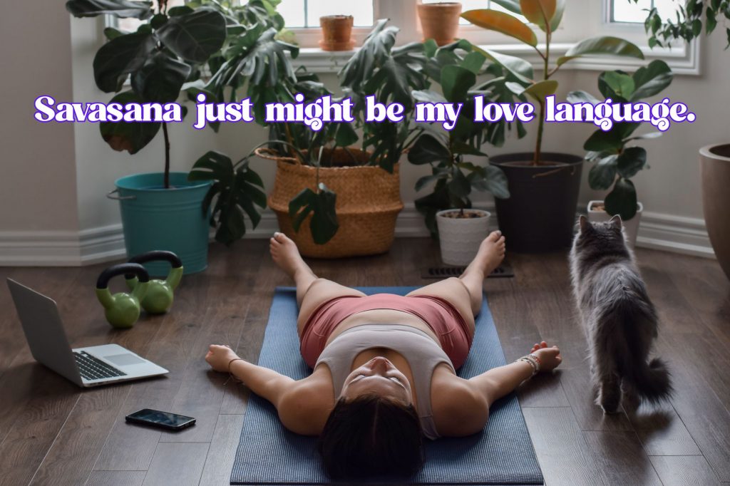 Woman lying in savasana on yoga mat on floor in a room full of plants, plus a laptop, kettle bells, a cell phone, and a cat. Text overlay reads "Savasana just might be my love language" 
